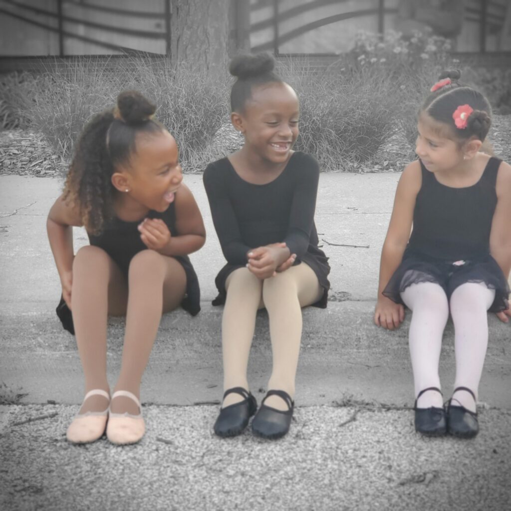 Three girls in ballet shoes and leotards sitting outside, smiling & laughing.